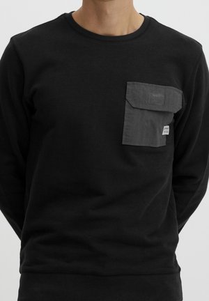 Man wearing black long-sleeve shirt featuring a dark gray rectangular chest pocket with a flap and small white label.