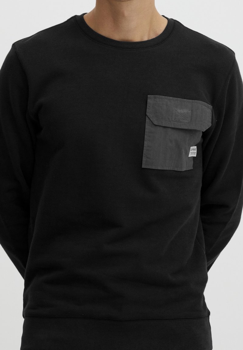 Homme portant une chemise noire à manches longues avec une poche poitrine rectangulaire gris foncé avec rabat et petite étiquette blanche.