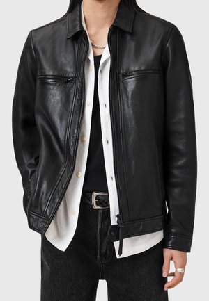 Personne portant une veste en cuir noire sur une chemise blanche déboutonnée et un haut noir, avec un jean noir et une bague en argent à la main droite.