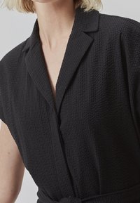 Vêtement en tissu texturé noir avec un col à revers, des manches courtes et une taille nouée. Présente un motif à carreaux subtil sur toute la surface.