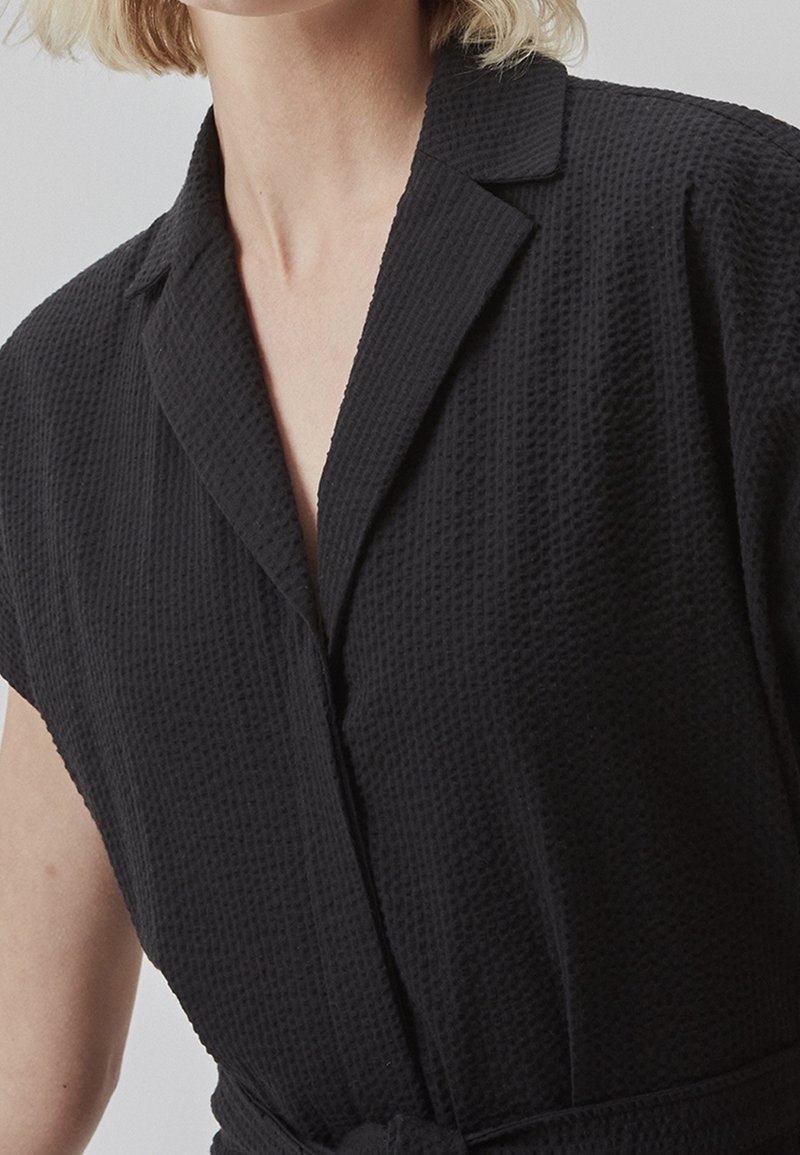 Vêtement en tissu texturé noir avec un col à revers, des manches courtes et une taille nouée. Présente un motif à carreaux subtil sur toute la surface.