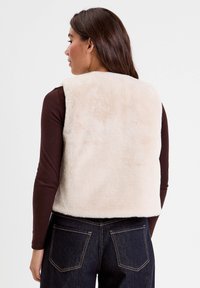 Femme vue de dos portant un gilet en fausse fourrure de couleur crème sur un haut à manches longues marron foncé et un jean bleu foncé avec des poches arrière visibles.