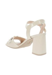 Sandalo con tacco in pelle verniciata beige, con punta aperta, cinturini incrociati e un robusto tacco a blocco. Presenta un fiocco decorativo e accessori in argento.