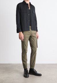Chemise noire boutonnée avec deux poches poitrine, associée à un pantalon cargo olive avec poches latérales et poignets élastiques, portée avec des chaussures noires.