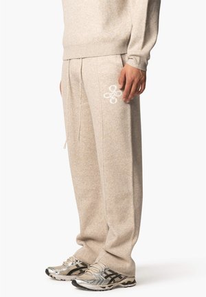 Beige Strick-Sweatpants mit elastischem Bund, Seitentaschen und einem weißen, gestickten Blumenmuster am linken Bein. Kombiniert mit silbernen Turnschuhen.