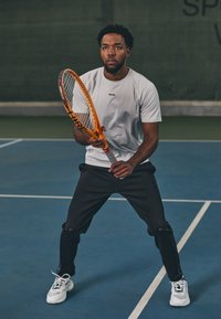 Tennisspelare i en ljusgrå skjorta och svarta byxor, som håller en orange tennisracket på en blå bana. Vita sneakers med svarta detaljer.