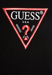 T-shirt en tissu noir avec un logo triangulaire rouge comportant le texte "GUESS U.S.A.", un point d'interrogation et "WASHED JEANS" en blanc autour.