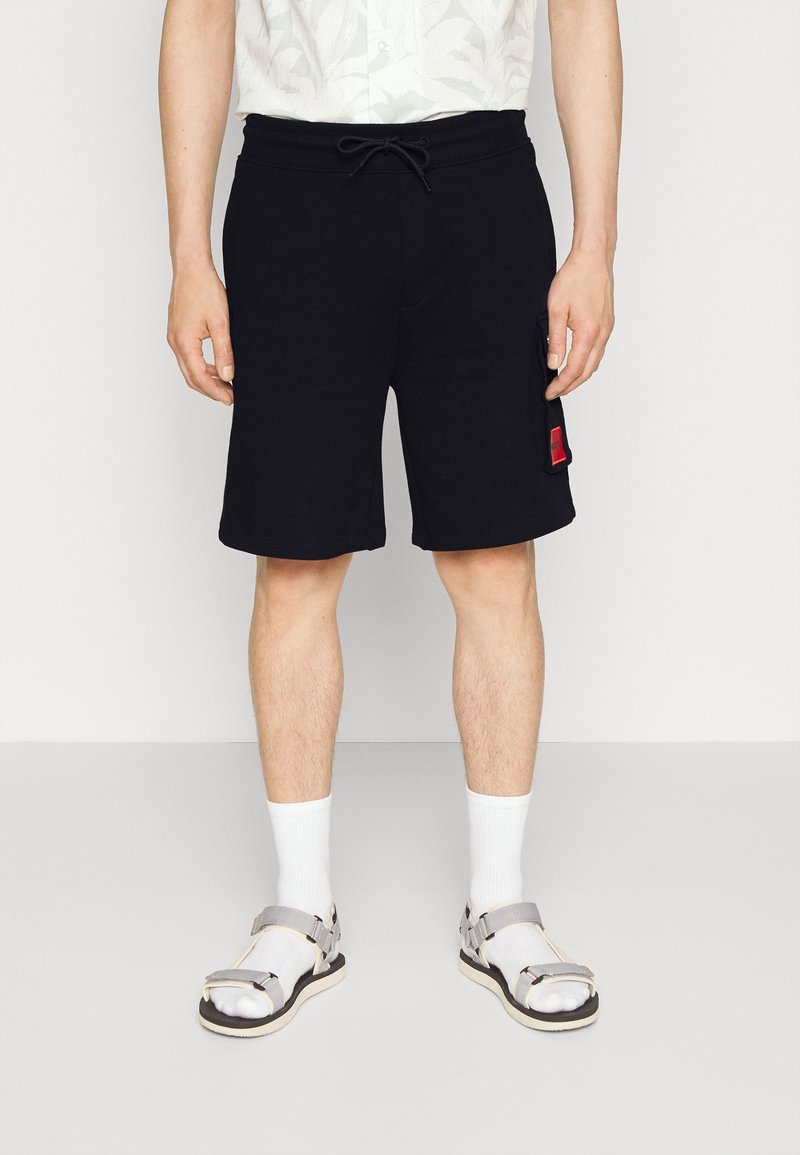 Shorts de algodón negros con cintura elástica y cordón ajustable, bolsillos laterales y una etiqueta de acento roja, combinados con calcetas blancas y sandalias grises.