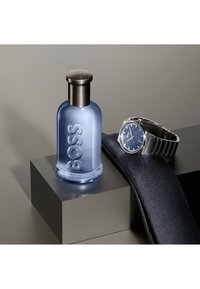 Blue Boss eau de cologne flaske med metal låg ved siden af sølvmetal armbåndsur på mørkeblå stof, vist på en grå piedestal.