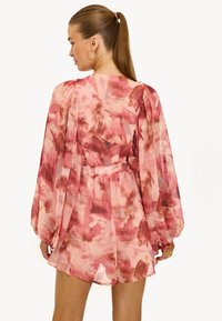 Vrouw met een lange paardenstaart, die grote oorbellen draagt en een roze-rode patroon lange jurk met pofmouwen aan heeft, gezien van achteren.