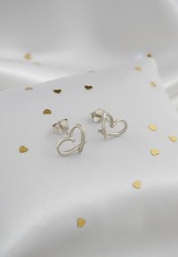 Silver hjärtformade studörhängen med en polerad finish, uppvisade på en vit satinkudde med spritt guldhjärt-konfetti.