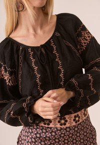 Blusa negra con escote fruncido y mangas largas, decorada con patrones bordados en rosa. Combinada con una falda de estampado floral.