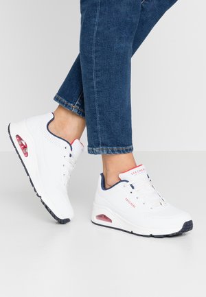 Osoba nosząca białe sneakersy Skechers z czerwonymi i granatowymi detalami oraz niebieskie dżinsy, stojąca na jasnoszarej podłodze i tle.