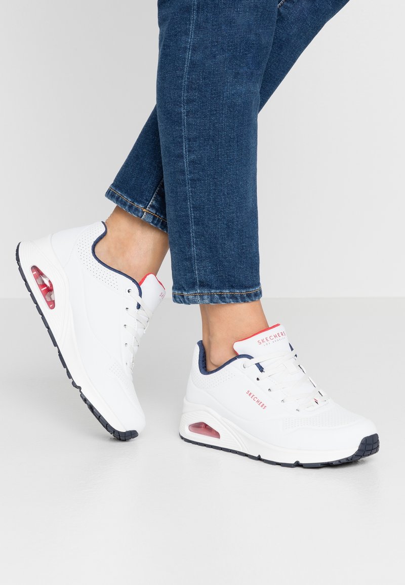 Personne portant des baskets blanches Skechers avec des détails rouges et bleu marine et un jean bleu, debout sur un sol et un fond gris clair.