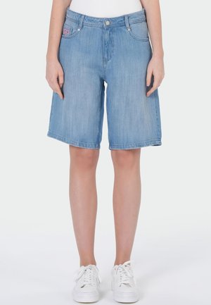 Hellblaue Denim-Knie-Shorts, getragen mit weißen Sneakern von einer Person, die vor einem einfarbigen Hintergrund steht.