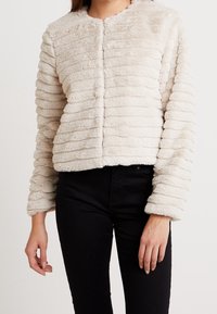 Texturerad, krämfärgad kort jacka med rund krage och horisontell ribbad design. Tillverkad av mjukt, plyschigt tyg; inga fästen synliga.