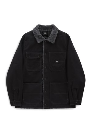 Homme portant une veste noire doublée sherpa, une chemise rose, un pantalon gris et des chaussures blanches sans lacets, debout avec une main dans la poche.