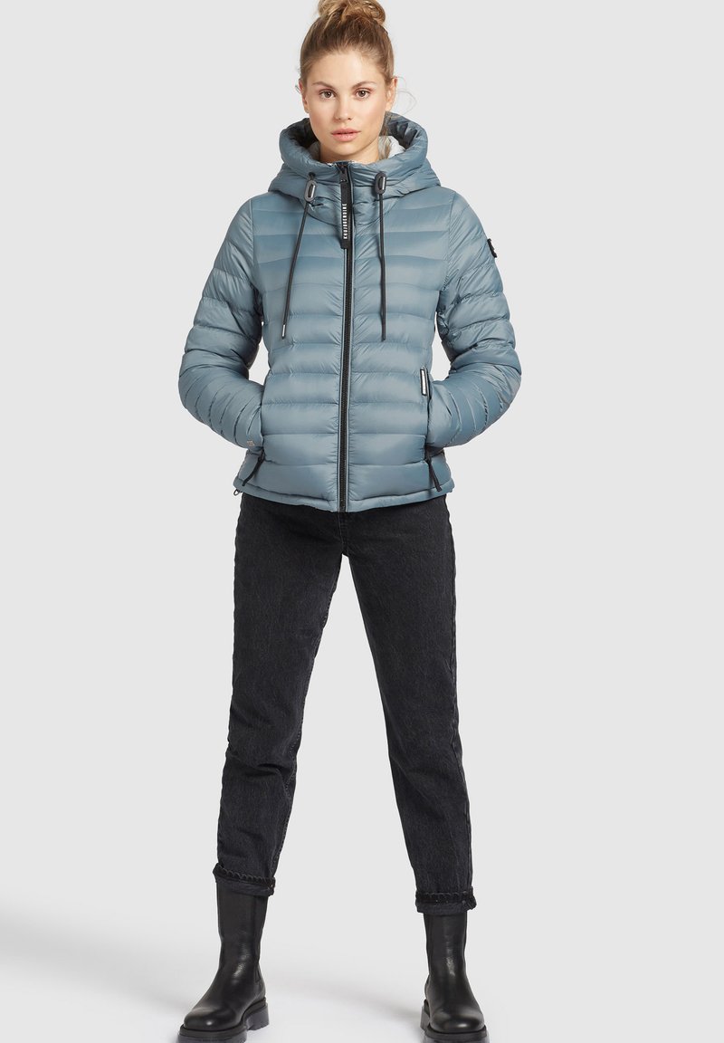 Helle blaue Daunenjacke mit Kapuze, ausgestattet mit einem Frontreißverschluss, verstellbaren Kordelzügen und Seitentaschen. Getragen mit schwarzen Jeans und Stiefeln.