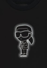 T-shirt noir avec un graphisme de dessin animé représentant une silhouette en lunettes de soleil et en costume, entourée d'un effet de halo. "KARL LAGERFELD" imprimé en dessous.