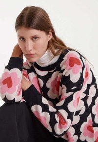 Mujer joven con cabello largo castaño que lleva un suéter negro con grandes patrones florales en rosa y rojo y un cuello de tortuga blanco debajo.