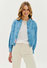 Světle modrá zkrácená bunda s vyšitými květinovými vzory, s kulatým límcem, zapínáním na knoflíky a elastickými manžetami. Nositá přes bílou košili.