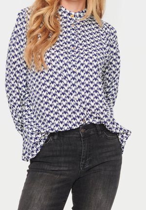 Frau trägt ein blau-weiß gemustertes, langärmliges Hemd mit Knöpfen, das in hoch taillierte schwarze Jeans gesteckt ist.