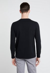 Långärmad svart skjorta i ett mjukt tyg, med rund hals och åtsittande ärmslut, ihop med ljusgrå byxor.