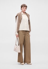 Μπεζ πλεκτό γιλέκο πάνω από ένα κρεμ πουκάμισο με κουμπιά, συνδυασμένο με καφέ παντελόνι φαρδύ και ανοιχτόχρωμες γόβες. Κρατάει μια μαλακή ροζ τσάντα.