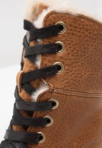 Bota de cuero marrón con cordones negros, ojales de latón y un forro de piel sintética en la parte superior. Superficie texturizada con detalles de costura.