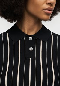 Γυναίκα που φοράει μαύρο πόλο πουκάμισο με λευκές κάθετες ρίγες και δύο κουμπιά, με εμφανές μέρος του λαιμού και της γραμμής του σαγονιού της.