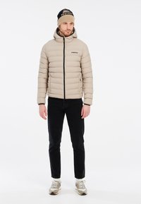 Veste doudoune beige avec des coutures horizontales, fermeture éclair noire et poches. Associée à un pantalon noir et un bonnet beige et noir.