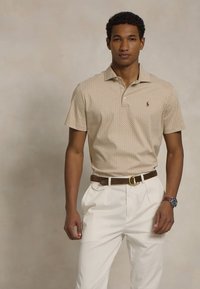Beige mønstrete poloskjorte med krage, kombinert med hvite plissert bukser og et brunt belte. Modell som bærer et klokke.