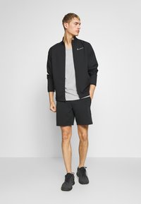 Giacca atletica nera con zip e maniche lunghe, t-shirt grigia sotto, pantaloni corti neri e scarpe atletiche nere. Design e vestibilità semplici.