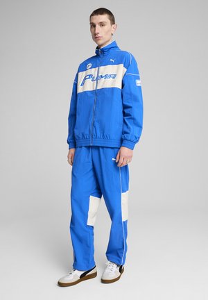 Ensemble de survêtement Puma bleu comprenant une veste zippée avec une bande horizontale blanche et un logo, assorti de pantalons et de baskets blanches.