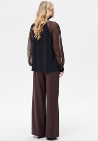 Czarna bluzka z przezroczystymi, wzorzystymi rękawami, w połączeniu z luźnymi brązowymi spodniami. Teksturowane detale na rękawach i miękkie wykończenie tkaniny na całej powierzchni.