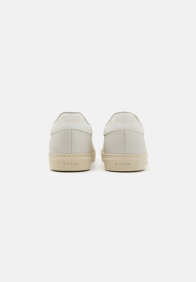 Zapatillas de cuero blancas con una textura suave, suela de goma crema y logo dorado en relieve en el talón. Diseño minimalista, forma redondeada.