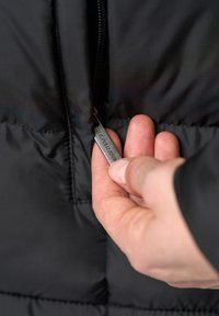 Juoda pūkinė striukė su lygiu paviršiumi ir blizgiu apdaila. Sidabrinė užtrauktuko su "U.S. POLO ASSN." įrašu trauka atidaro kišenę.