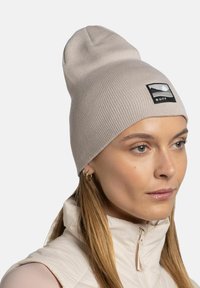 Beige knit beanie with a rounded top and a black rectangular patch featuring a mountain silhouette and "BUFF" text. Smooth texture.