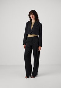 Schwarzer taillierter cropped Blazer über einem beigen Top, kombiniert mit locker sitzenden schwarzen Hosen und offenen schwarzen Sandalen. Glatte Texturen, minimales Design.