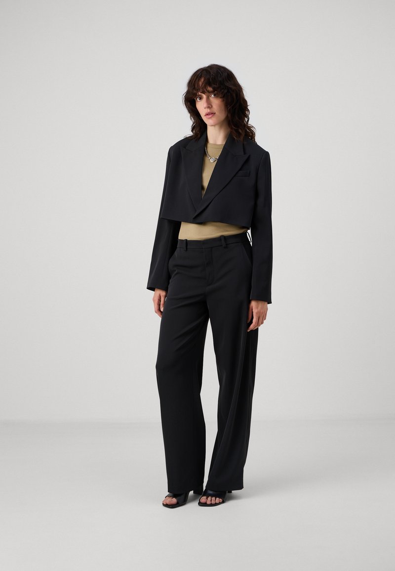 Zwarte op maat gemaakte cropped blazer over een beige top, gecombineerd met losse zwarte broeken en open-teed zwarte sandalen. Glad texturen, minimalistisch ontwerp.
