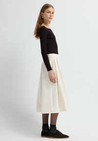 Haut noir à manches longues côtelé associé à une jupe midi plissée crème. Le modèle porte des chaussures sombres et des chaussettes grises, se tenant en profil sur un fond clair.