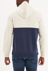 Sudadera con capucha bicolor en crema y marino, con tejido suave, bolsillo frontal, dobladillo y puños acanalados. Detalles de costura visibles.