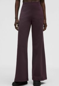 Osoba oblečená v tmavo purpurových nohaviciach s vysokým pásom a širokými nohavicami, s prednými švami a bočnými vreckami na zips, zasadená do čiernych topánok, na bielom pozadí.