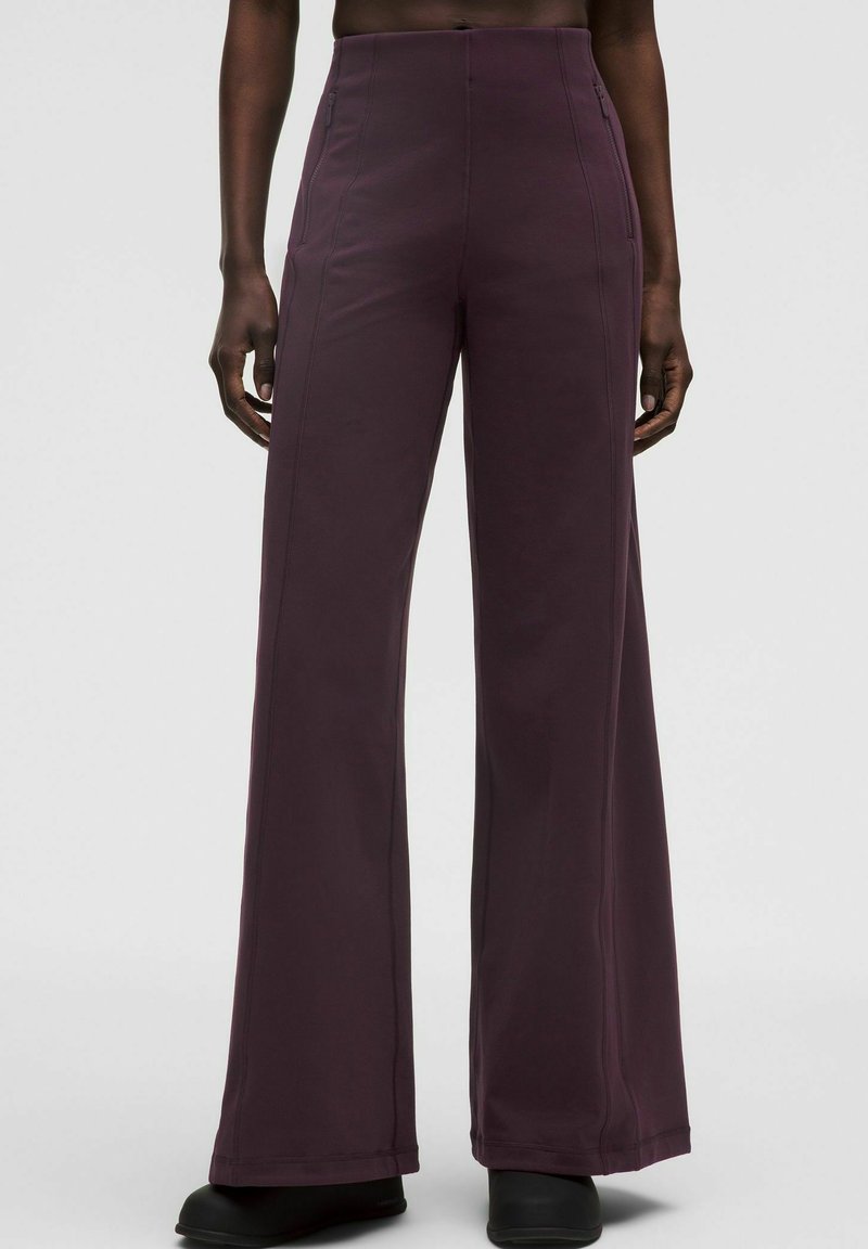 Osoba oblečená v tmavo purpurových nohaviciach s vysokým pásom a širokými nohavicami, s prednými švami a bočnými vreckami na zips, zasadená do čiernych topánok, na bielom pozadí.