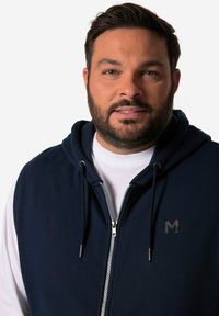 Navy zip-up hoodie with a front pocket, drawstrings, and a logo accent. The fabric appears soft and textured, suitable for casual wear.