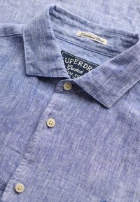 Chemise boutonnée bleu clair texturée avec col ouvert, boutons blancs et étiquettes de marque visibles à l'intérieur de l'encolure.