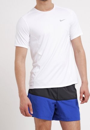 Mann trägt ein weißes Nike-Sportshirt mit kurzen Ärmeln und blaue und schwarze Shorts, steht mit entspannten Armen an den Seiten.