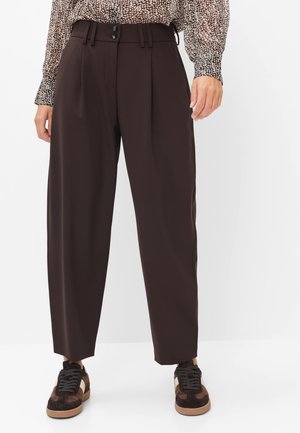 Vrouw met donkerbruine wijde broek met hoge taille, een bedrukte blouse die is ingestoken en bruine en zwarte vetersneakers.
