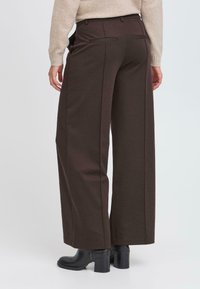 Pantalons marron à jambes larges en tissu texturé, dotés de poches arrière fines et d'une taille haute, accompagnés de bottines noires à talons.