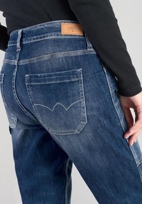 Persona que lleva puestos unos pantalones vaqueros de denim desgastados de color azul con bolsillos traseros y una etiqueta de marca de cuero marrón, vestida con una camiseta de manga larga negra.